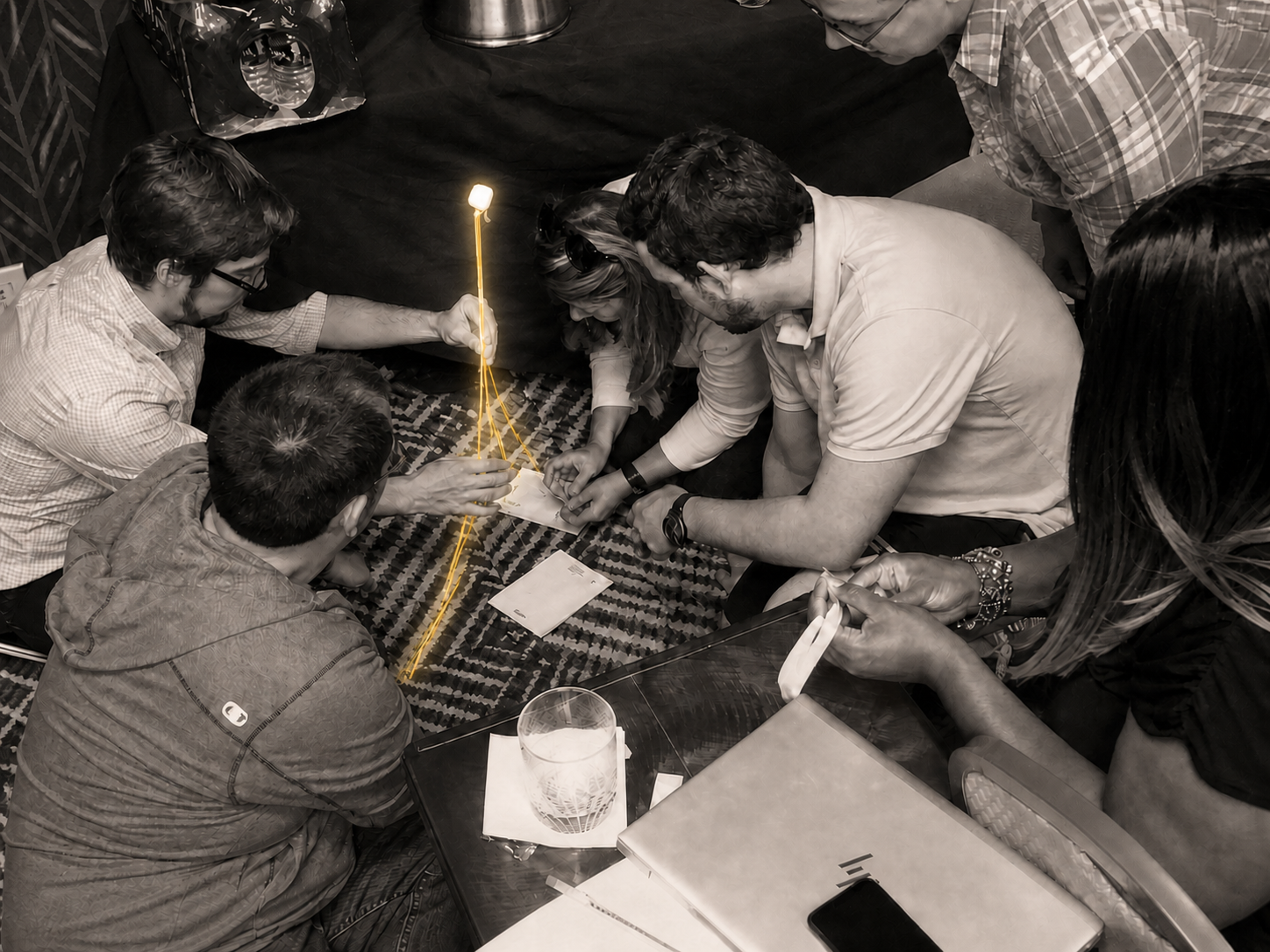 A team in warm monochrome collaborating on a glowing spaghetti-and-marshmallow structure